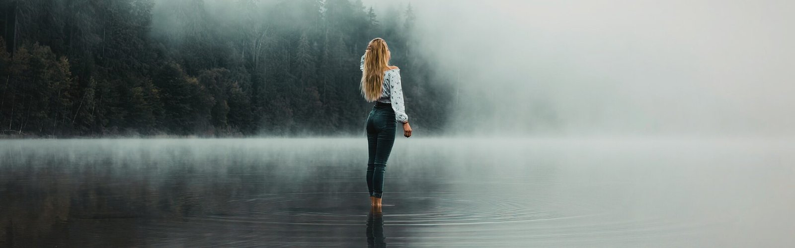 Personne seule debout sur un lac calme, regardant la brume et les arbres à l'horizon, symbolisant le retrait et la contemplation pendant un burn-out.