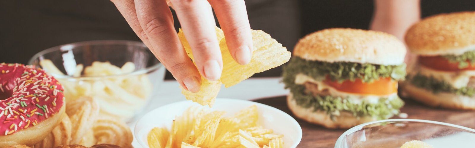 Une main saisit une chips au-dessus d’un bol, entouré de différents aliments tels que des burgers, des donuts et des chips, illustrant un moment de prise alimentaire impulsive.