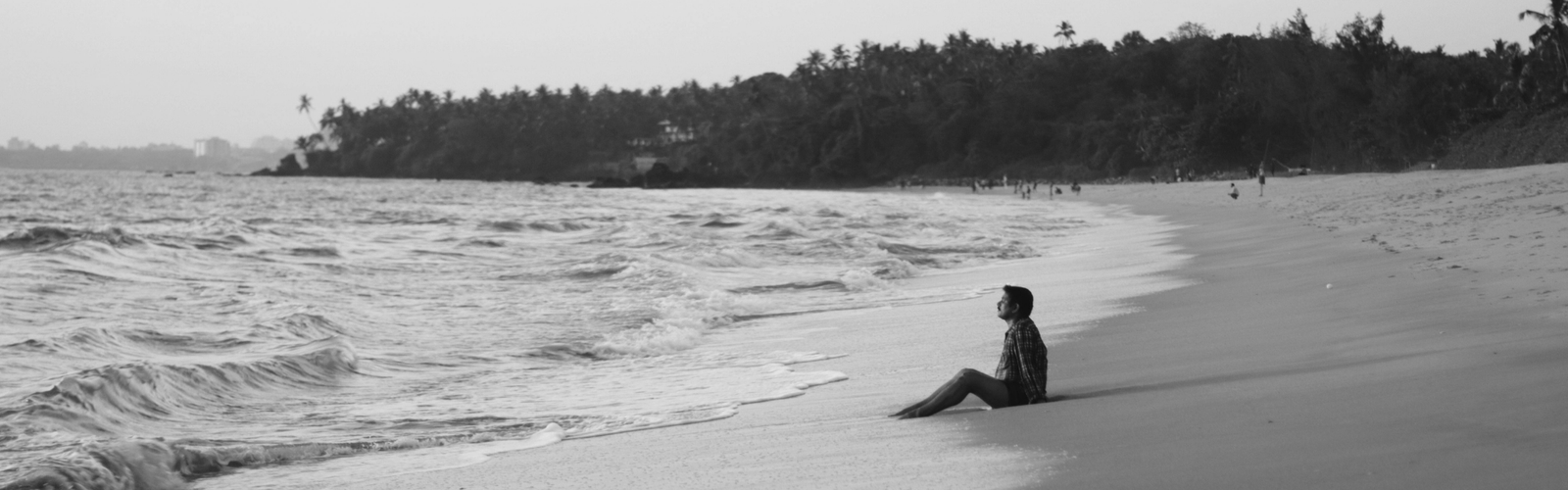 Personne assise sur une plage face à la mer, tandis que d’autres personnes se trouvent au loin.
